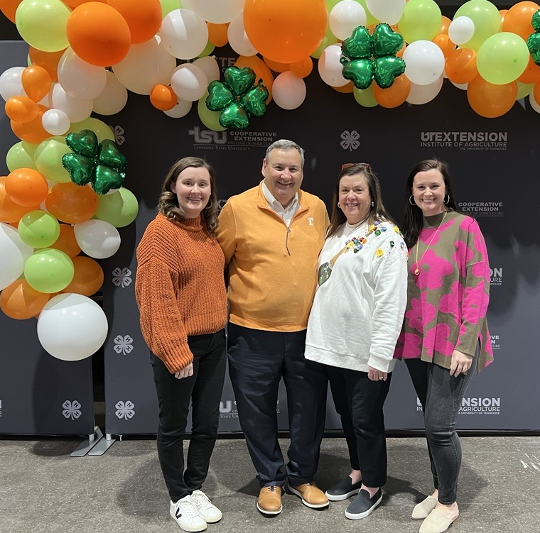 Rachel Smith with two other women and a man in front of a UT Extension and 4-H backdrop with orange, white, and green balloons