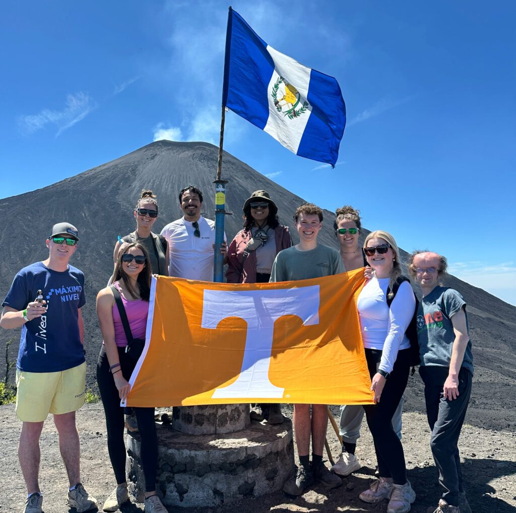 Kyra Barrier is among a group of people standing on a rocky terrain with a distant volcano in the background. They are holding a large orange flag with the University of Tennessee logo. Above them, the national flag of Nicaragua is visible.