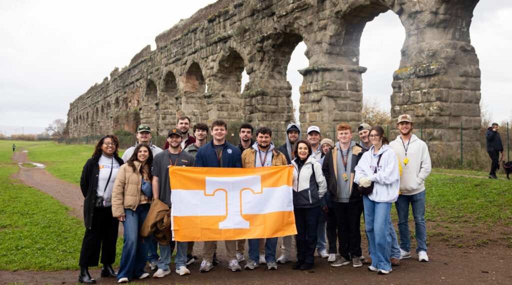 Kyra Barrier among a group of students standing in front of the ancient aqueducts in Rome, Italy, holding an orange UT flag.