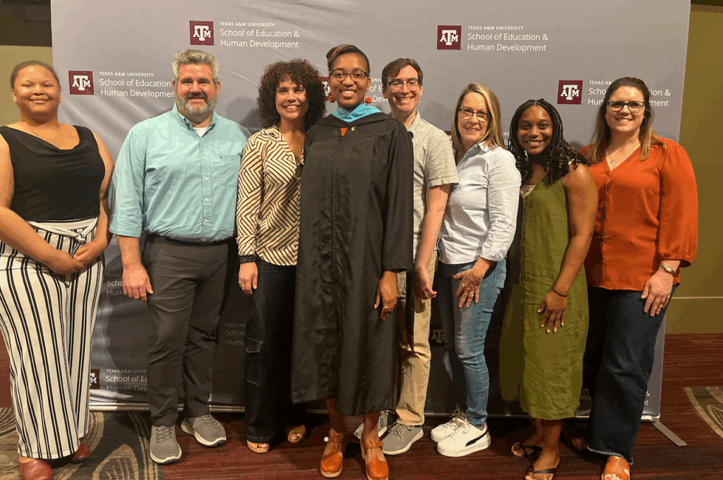 Kyra Barrier in a graduation gown standing at the center of a group in front of a sign with the Texas A&M School of Education and Human Development logo