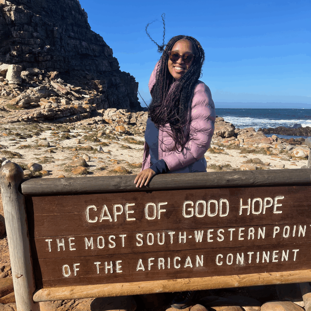 Kyra Barrier standing on a rocky beach with a sign that reads "Cape of Good Hope, the Most South-Western Point of the African Continent."