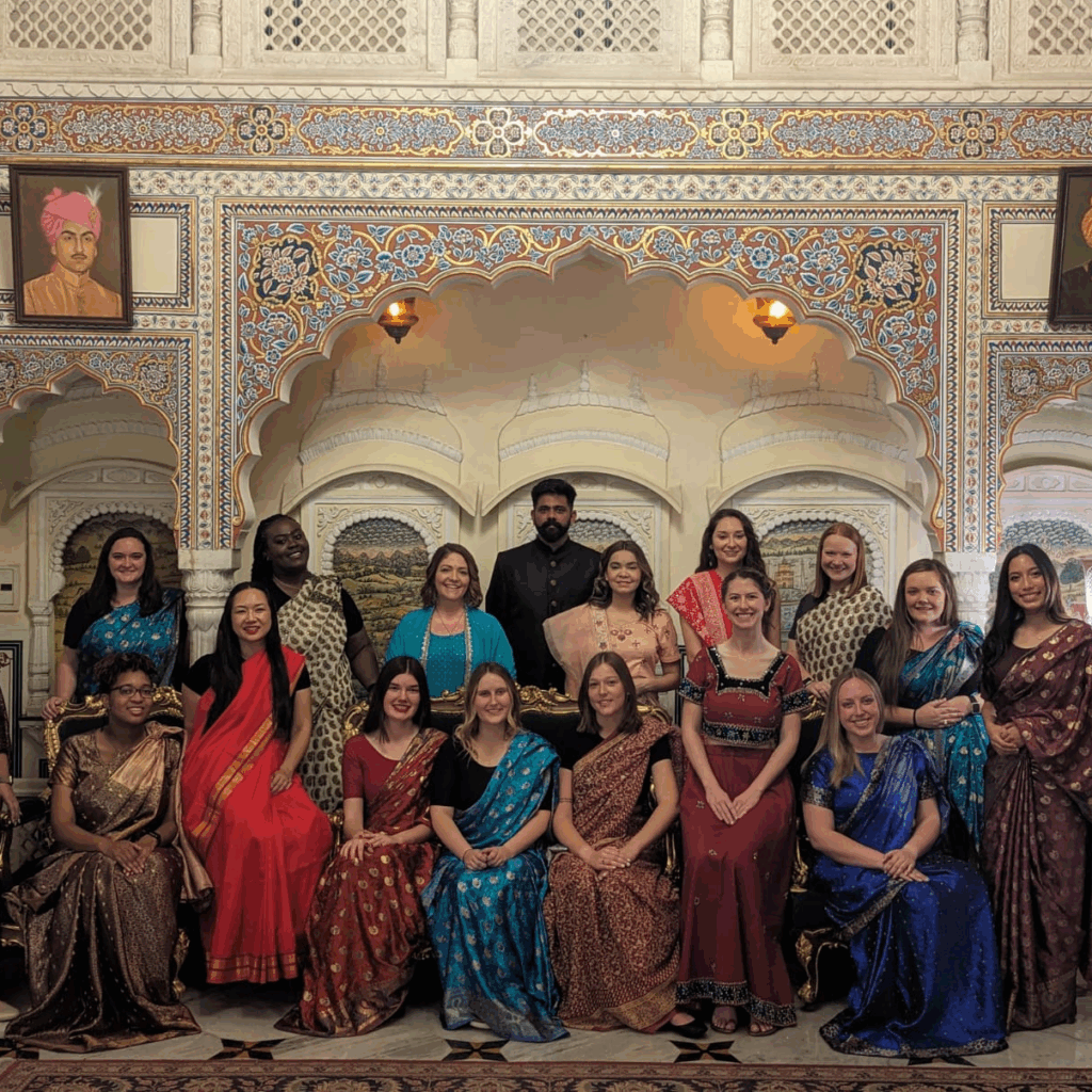 Kyra Barrier among a group of people dressed in colorful traditional Indian attire, posing in an ornately decorated room with patterned arches. The room features portraits of historical figures on the walls.