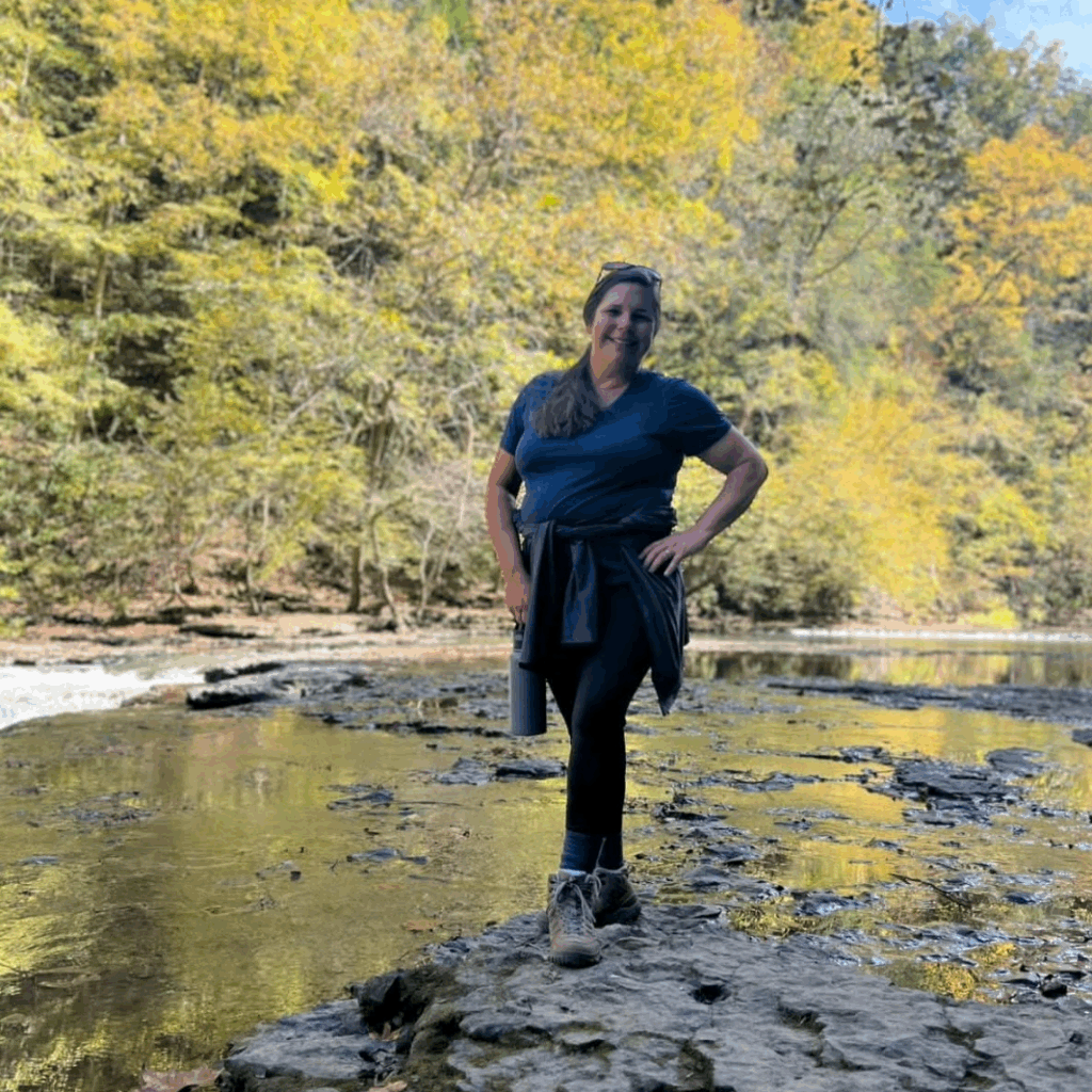 Shalena Durkot on a hike standing on the bank of a creek with colorful trees in the background.