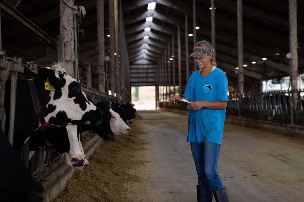 Mason Dunlap stands inside a spacious barn, holding a notebook and pen. She is in front of a few cows eating from a trough. The barn has a high ceiling and rows of lights.