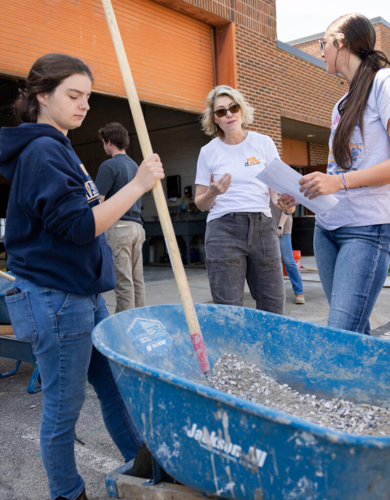 Angela Smith instructs two students during a concrete mixing lab. One student mixes gravel in a blue wheelbarrow with a long-handled tool.