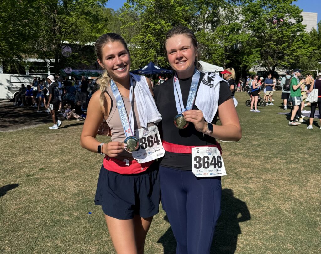 Mason Dunlap and a friend stand on a grassy area after completing a race, each holding a medal and wearing towels around their necks. A crowd is gathered in the background under a clear blue sky.