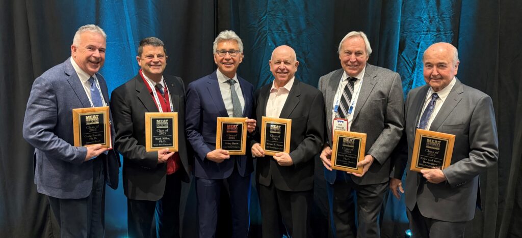 A group of six people, including Craig Bacon, is standing side by side, each holding a plaque with a black background and gold borders. They are dressed in formal attire, with suits and ties. A blue curtain is in the background.