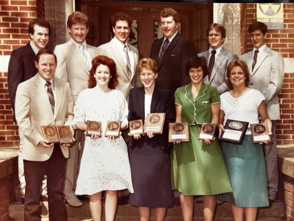 Eleven people stand in front of a brick building, posing with award plaques. They are dressed in formal attire and smile at the camera.