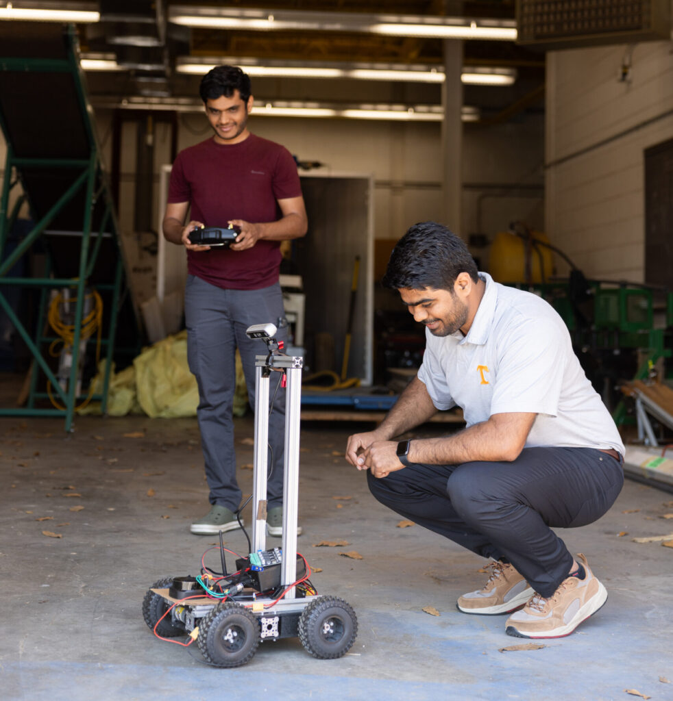 Chetan Badgujar and a student in a garage-like setting are engaging with a small wheeled robot on the floor. Badgujar is crouching beside the robot, which has visible electronics and wiring. The student stands, holding a remote control.