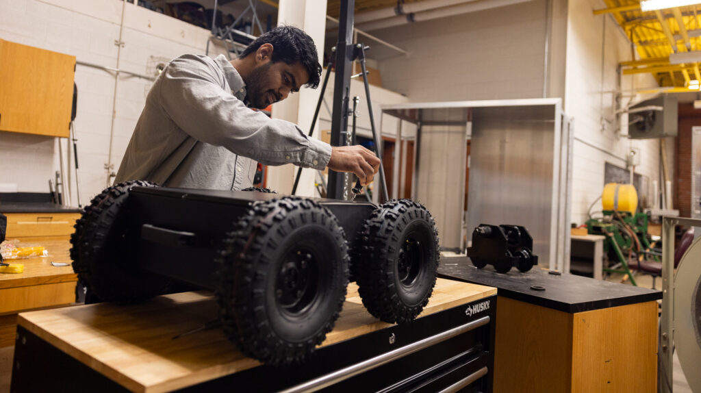 Chetan Badgujar working on a large wheeled robot on a workbench in a workshop. The robot has four large, rugged wheels.