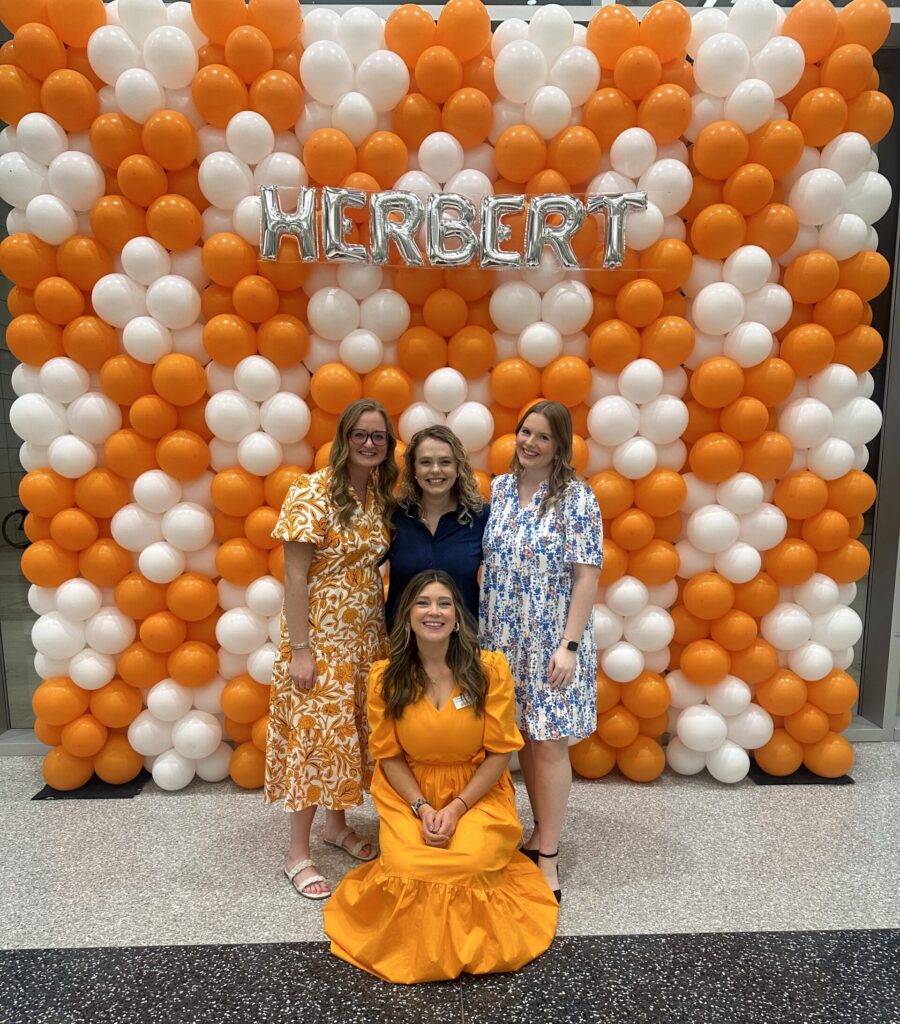 A group of people posing in front of a balloon backdrop with an orange and white color scheme. The backdrop has the name "Herbert" in metallic letters.