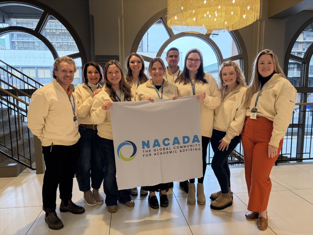Nine smiling individuals stand indoors holding a banner reading "NACADA: The Global Community for Academic Advising." They are in a well-lit area with large arched windows in the background.