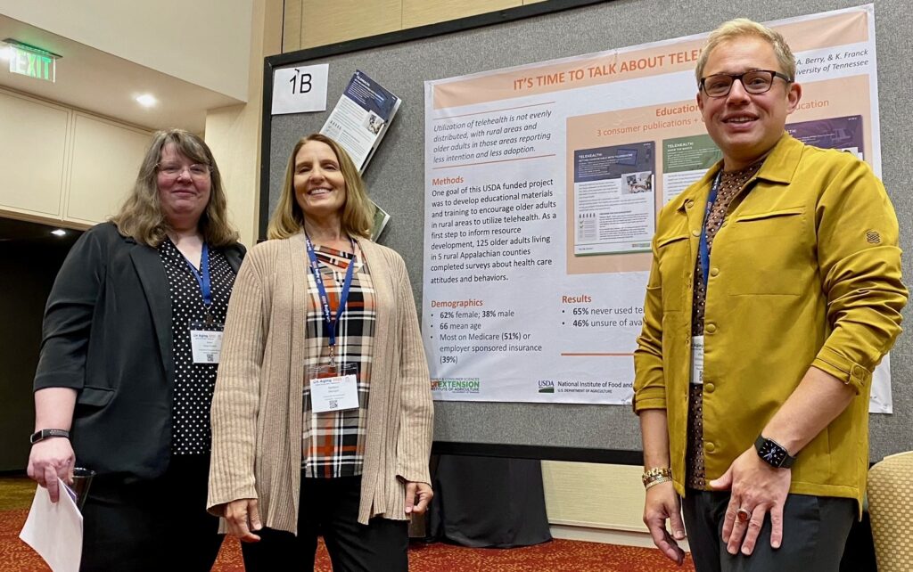 A group of three people standing in front of a presentation board at a conference. The presentation is titled "It's Time to Talk about Telehealth." One person is holding a stack of papers. All are smiling and wearing conference badges.