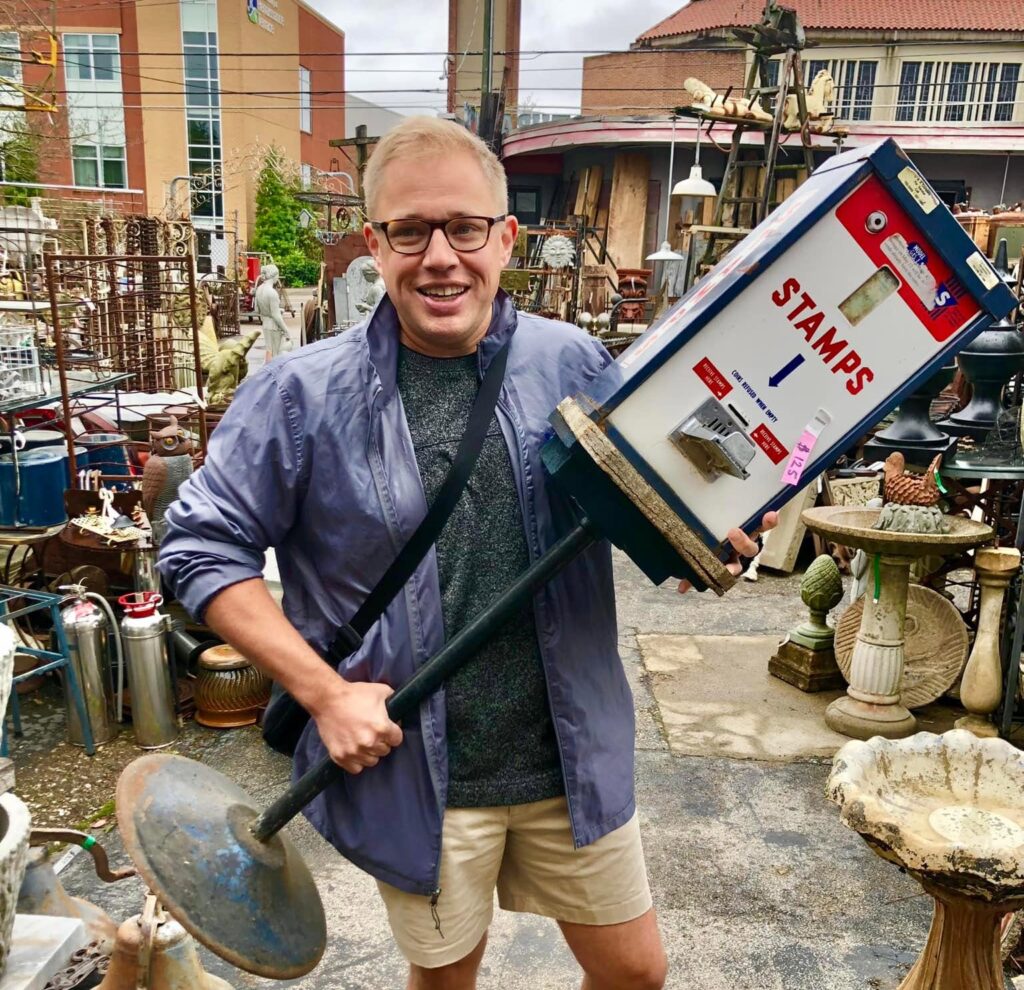Christopher Sneed stands outdoors at an antique market, smiling while holding a tall vintage USPS stamp dispenser.