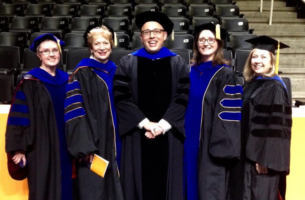 A group of five people wearing academic regalia, including caps and gowns, stand together smiling for a photo. They are indoors, with empty stadium seating visible in the background.