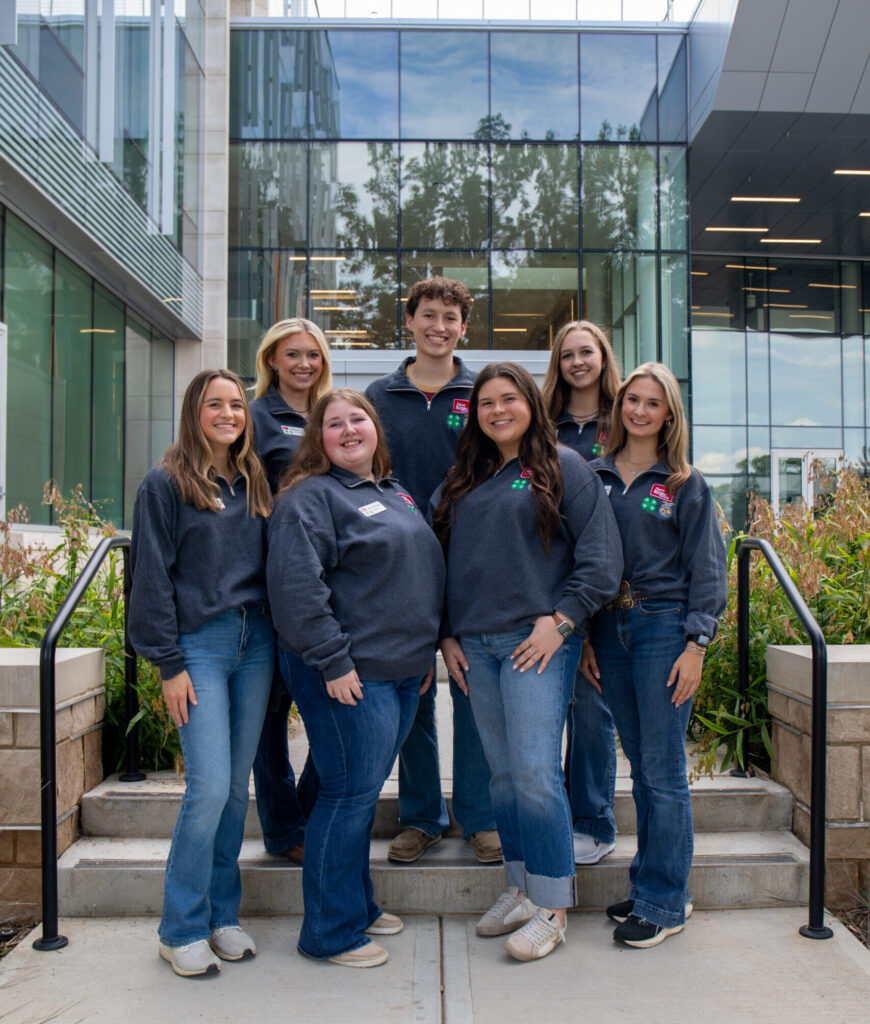 A group of seven people standing on steps outside the Agriculture and Natural Resources Building. They are all wearing matching gray tops with Farm Bureau, 4-H, and FFA logos