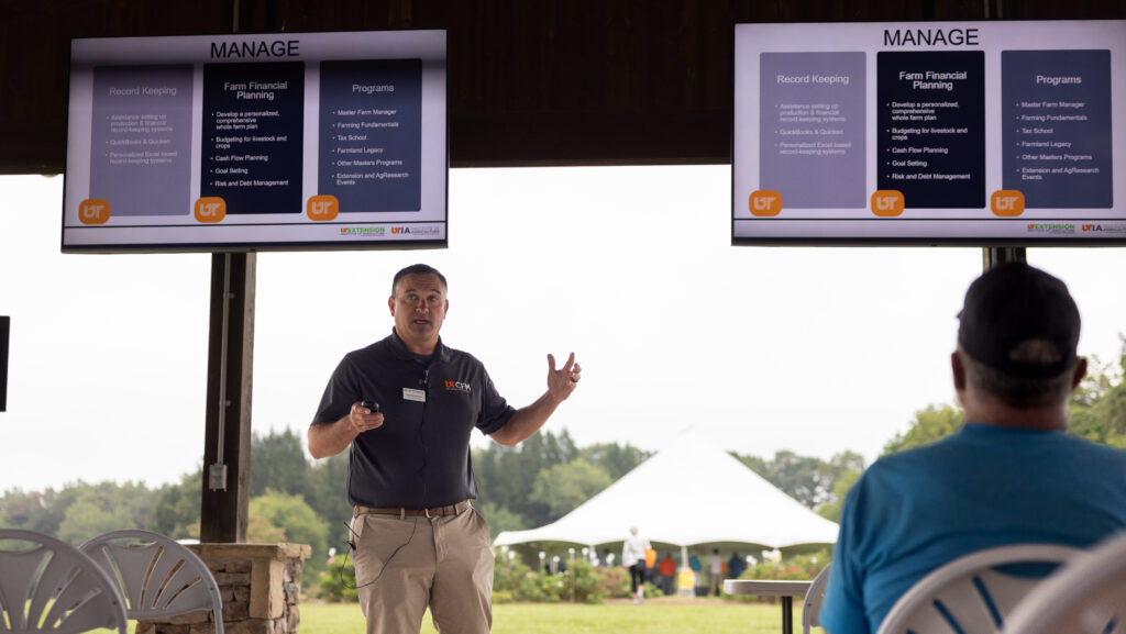 David Bilderback gives a presentation outdoors under a covered area with two screens displaying slides titled “MANAGE.” The slides cover topics related to financial planning and record-keeping.