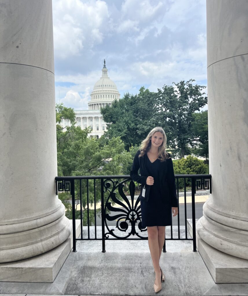 Skylar Baird stands on a balcony with the U.S. Capitol building visible in the background. She is dressed in formal attire, and the scene includes ornate railing and large columns.