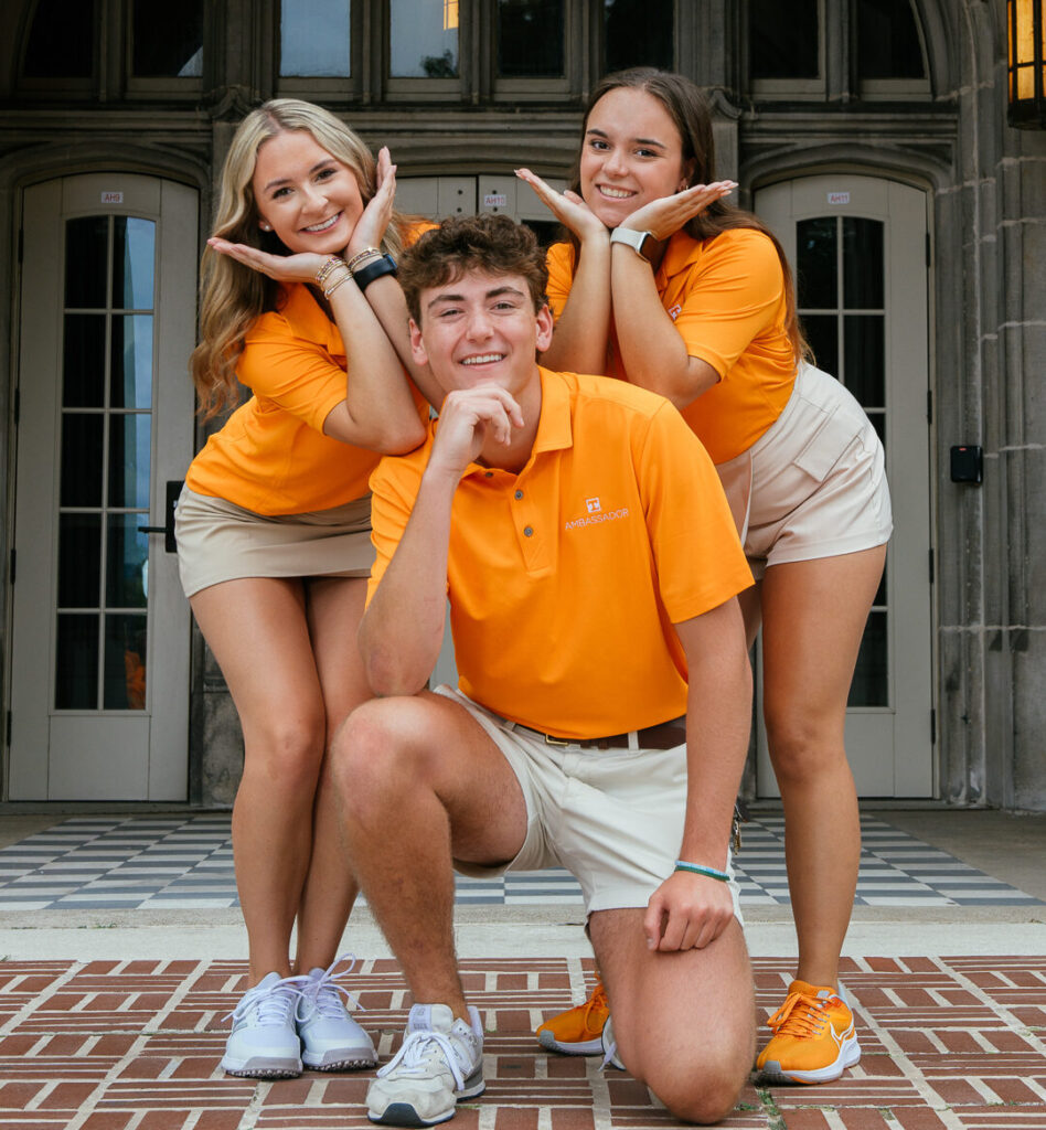 Three individuals wearing orange shirts and light-colored shorts pose playfully in front of an arched entrance with decorative stonework. Two stand behind with their hands under their chins, while one kneels in front on a brick pathway.