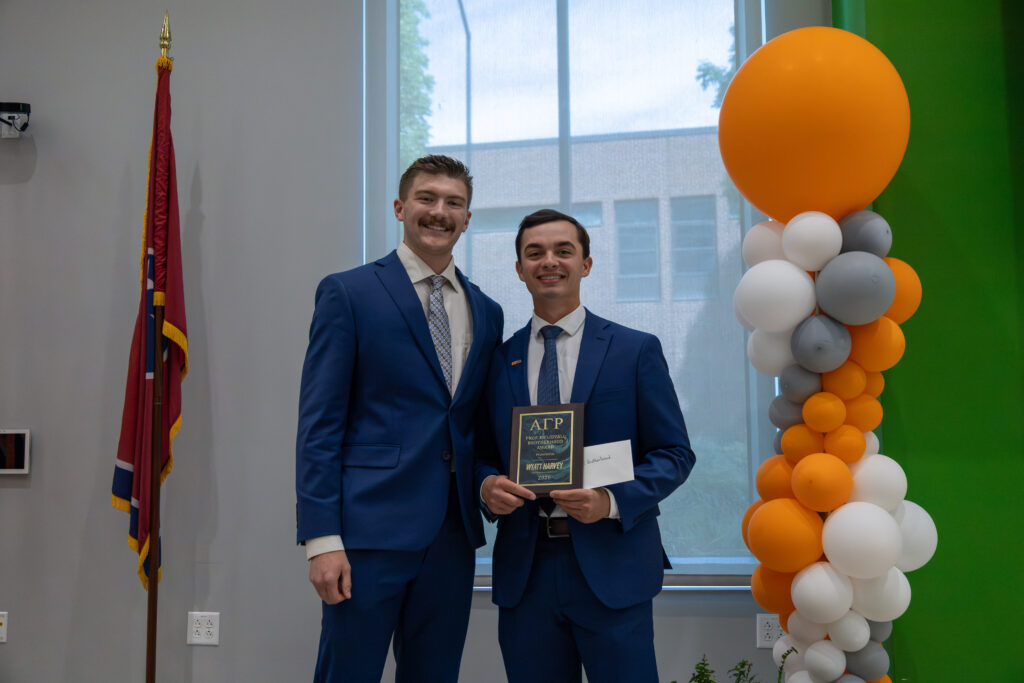 Two individuals are posing indoors, one holding a plaque with "AGR - Prof. Ed Lidvall Brotherhood Award Presented to Wyatt Harvey, 2026" on it. They are standing beside an orange and white balloon arrangement and a Tennessee state flag.