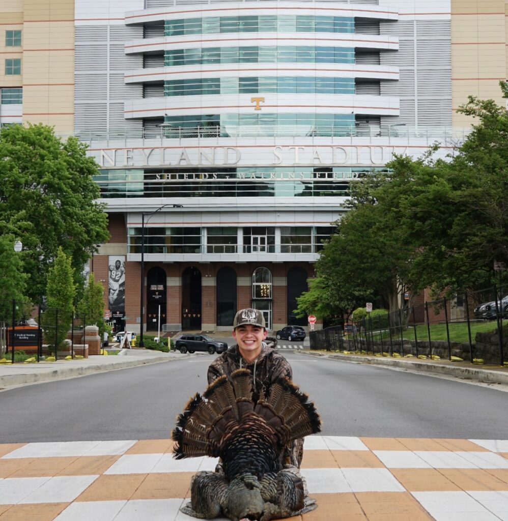 Wyatt Harvey kneels on an orange-and-white checkerboard crosswalk in front of Neyland Stadium, holding a large turkey.