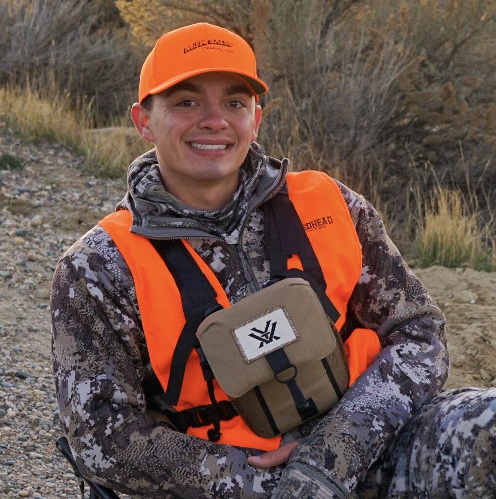Wyatt Harvey wearing an orange cap and vest, with camouflage clothing, sits outdoors on a portable chair.