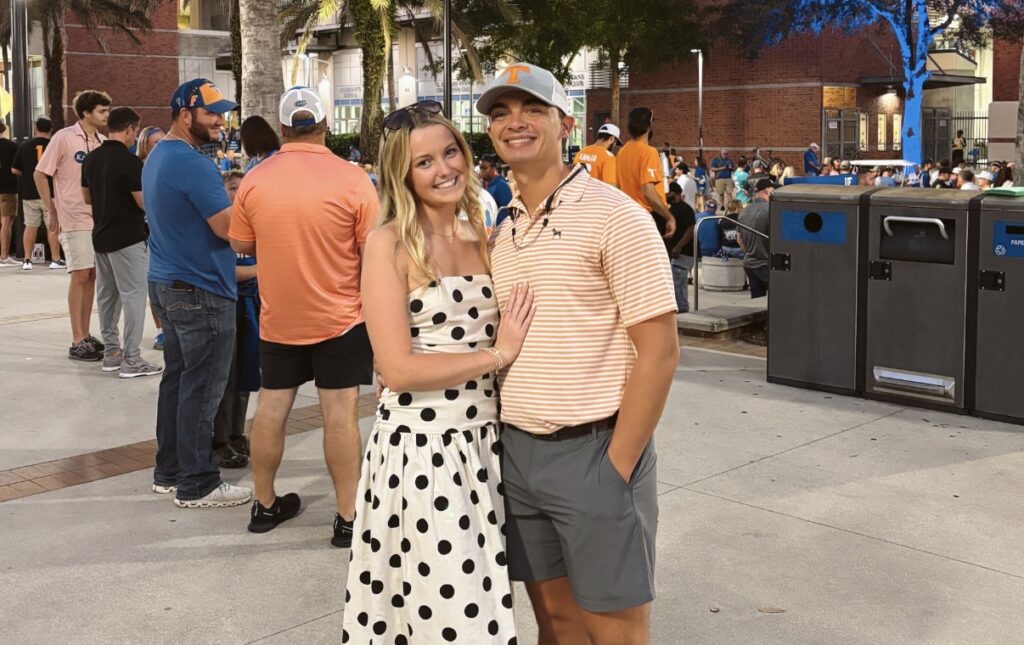 Wyatt Harvey and his girlfriend, who is wearing a black and white polkadot dress, smiling for a picture outdoors with palm trees in the background.