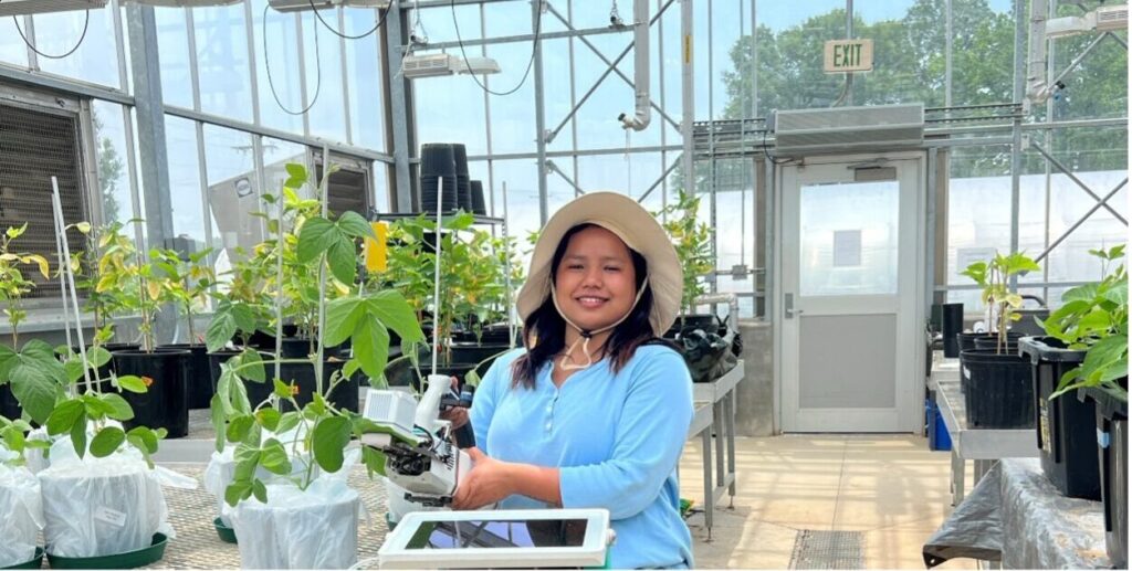 Smirti Shahi in a greenhouse setting, surrounded by plants and technical equipment. She is wearing a wide-brimmed hat and holding a tablet. The environment is well-lit with several large windows and transparent roofing.