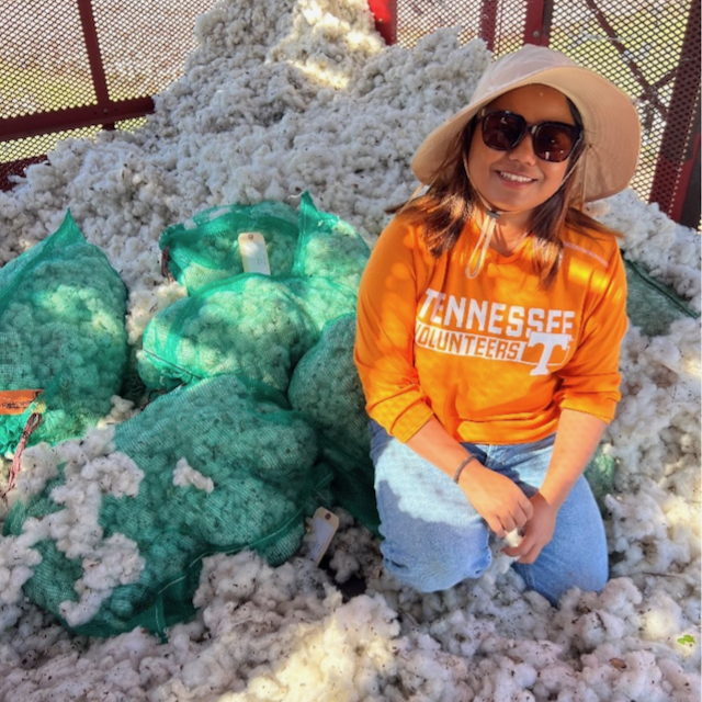 Smirti Shahi kneeling on a large pile of cotton, wearing a hat, sunglasses, and an orange "Tennessee Volunteers" shirt.