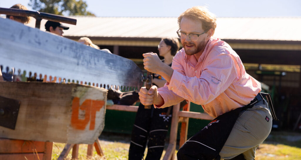 Max Brabson is engaging in a log sawing activity using a large saw. They are outdoors, and other participants are partially visible in the background.