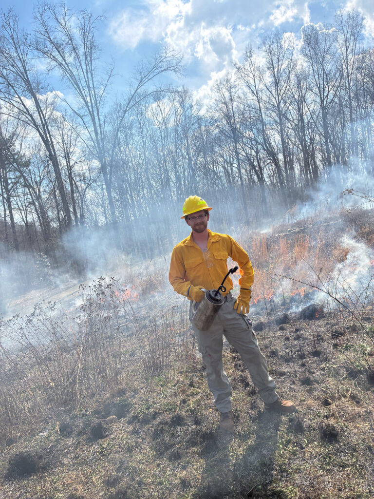 A person in protective clothing and a yellow hard hat stands in a field with patches of fire and smoke. The sky is partly cloudy, and trees without leaves are visible in the background. The person is holding a fire-setting tool.