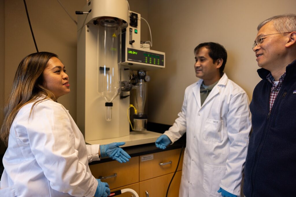 Qixin Zhong standing with two students wearing white lab coats in a lab