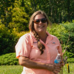 Portrait of Shalena Durkot smiling in a green garden setting, She is wearing sunglasses and is holding garden shears.