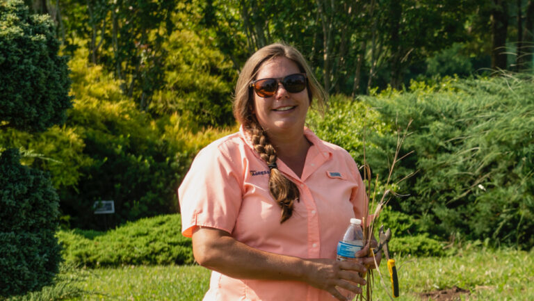 Portrait of Shalena Durkot smiling in a green garden setting, She is wearing sunglasses and is holding garden shears.