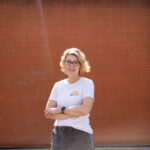 Angela Smith smiling in front of an orange garage door. She wears a white shirt featuring the words "BESS is Best."