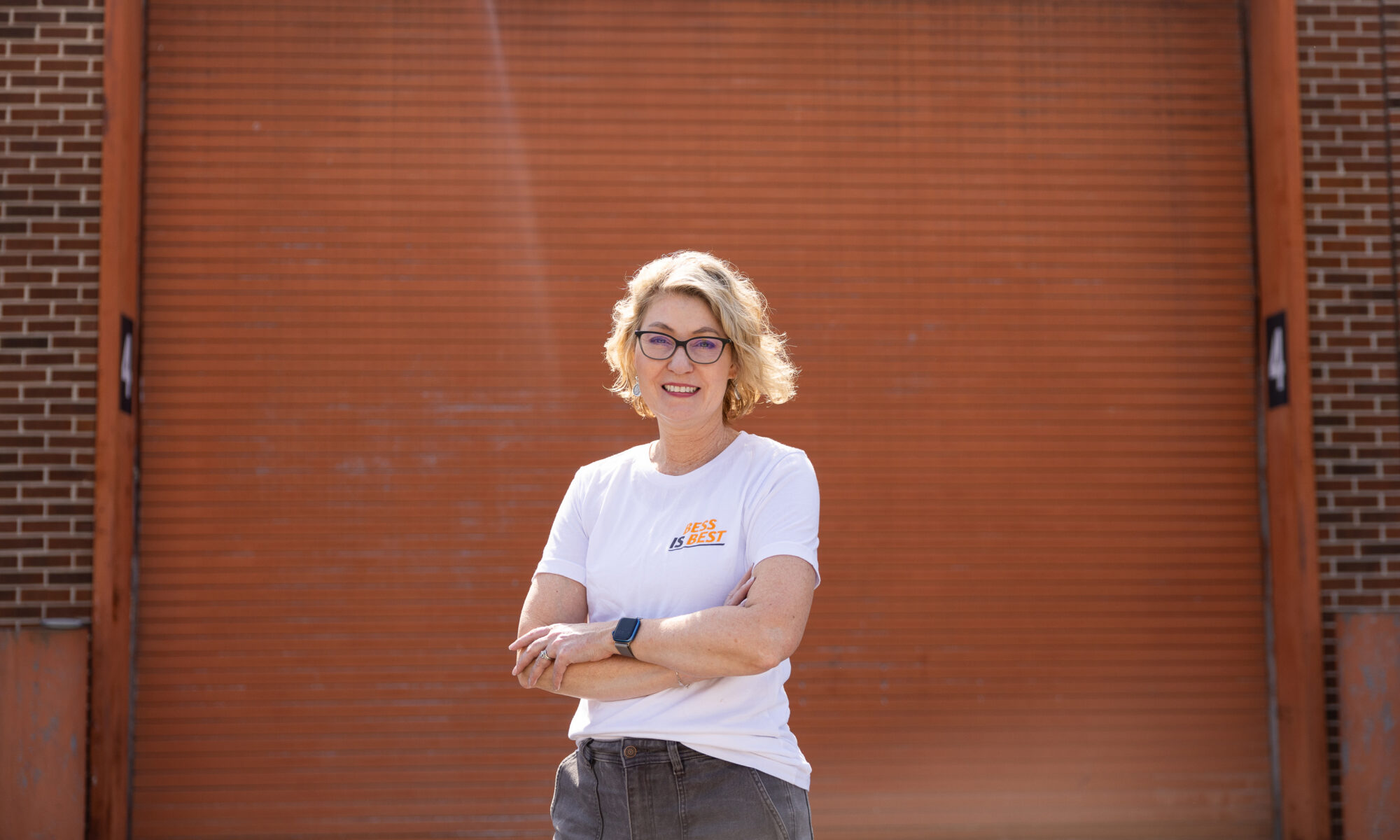 Angela Smith smiling in front of an orange garage door. She wears a white shirt featuring the words "BESS is Best."