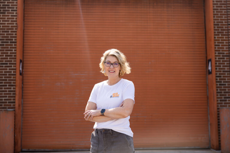 Angela Smith smiling in front of an orange garage door. She wears a white shirt featuring the words "BESS is Best."