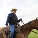 Kevin Thompson wearing a cowboy hat and a vest is riding a brown horse in a grassy field.