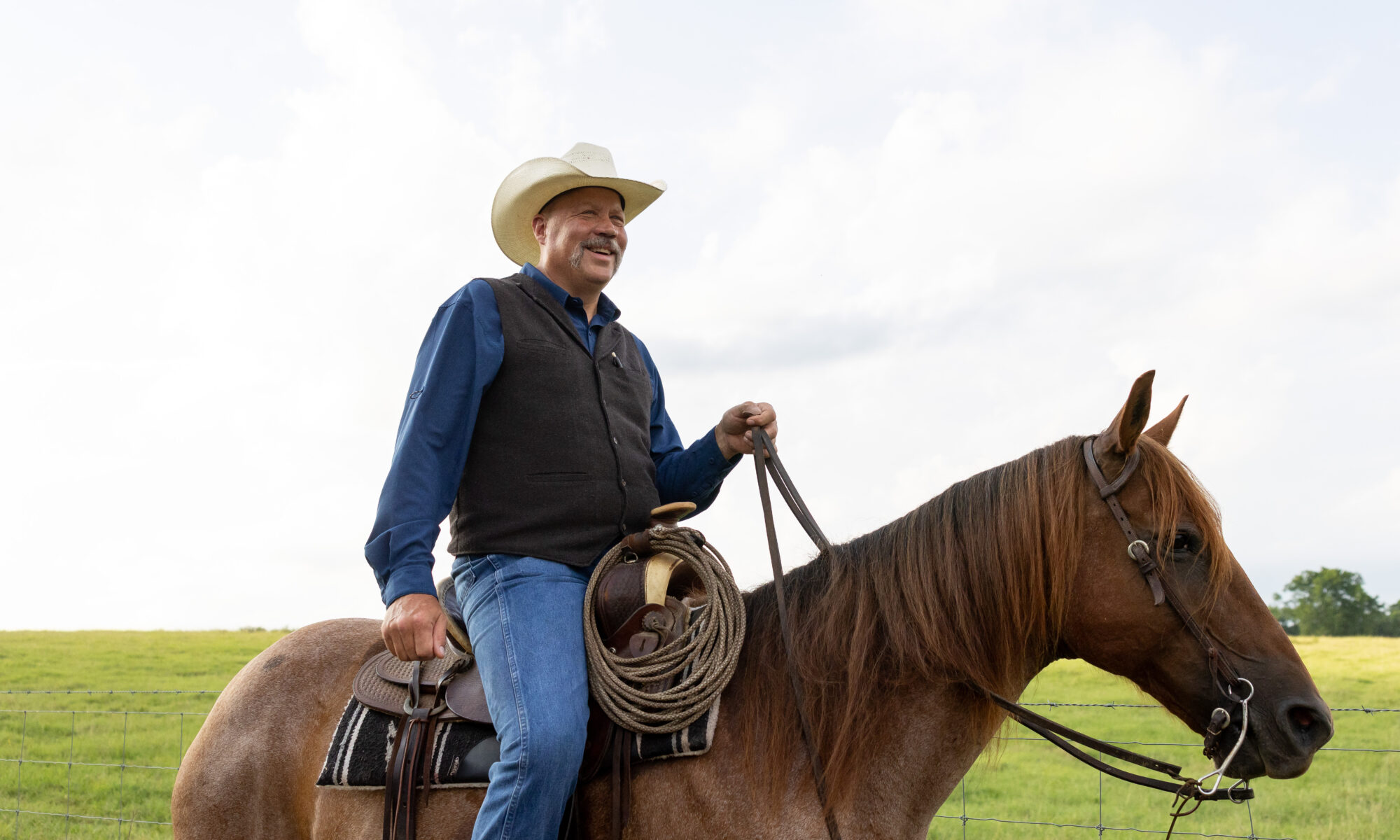Kevin Thompson wearing a cowboy hat and a vest is riding a brown horse in a grassy field.