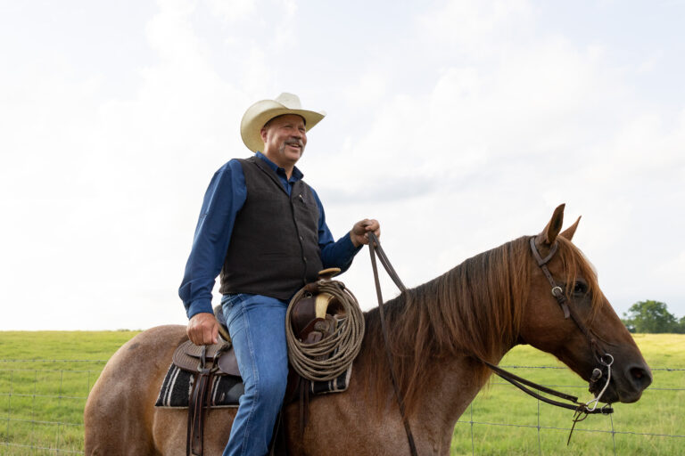 Kevin Thompson wearing a cowboy hat and a vest is riding a brown horse in a grassy field.