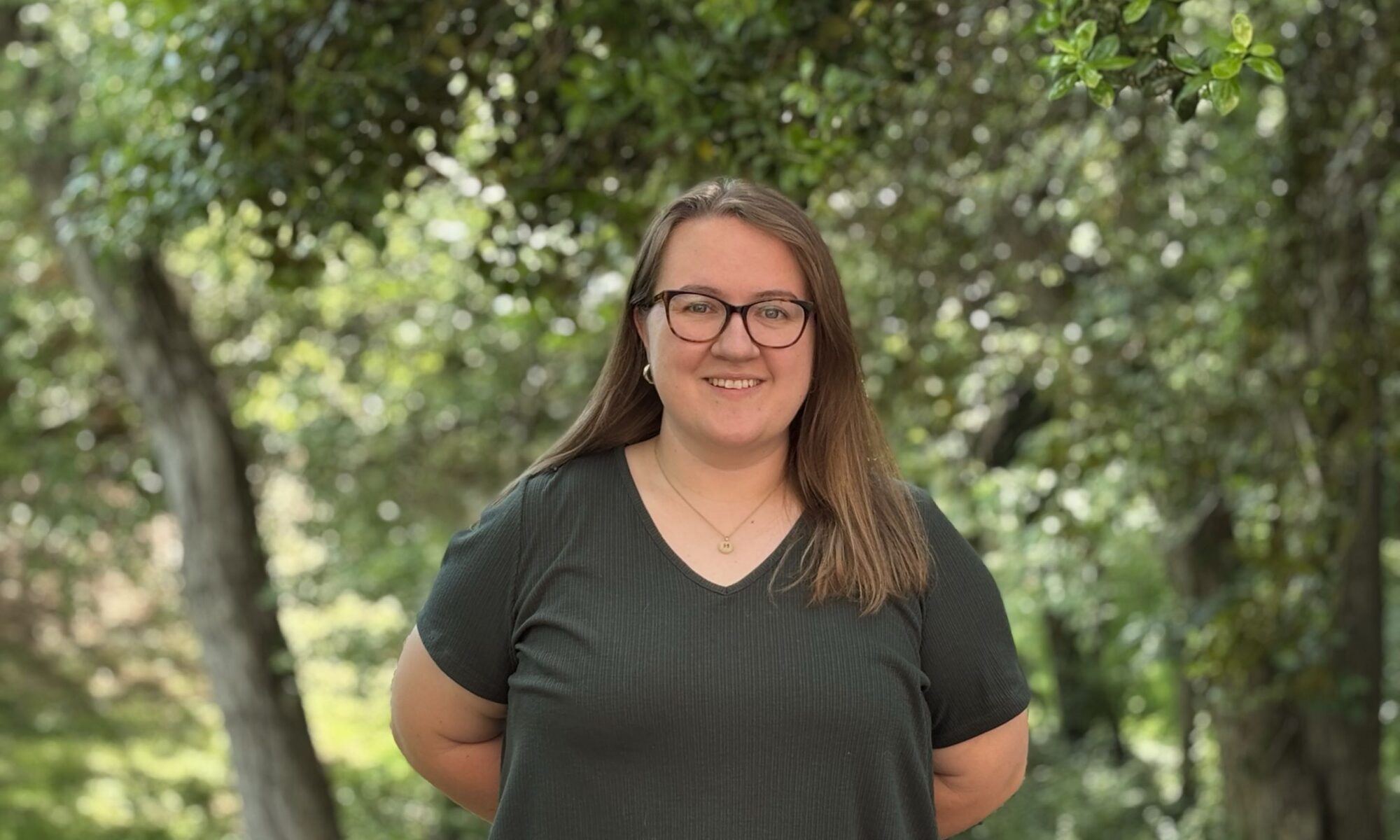 Haley Williamson in a green top smiles at the camera. Green trees are in the background.