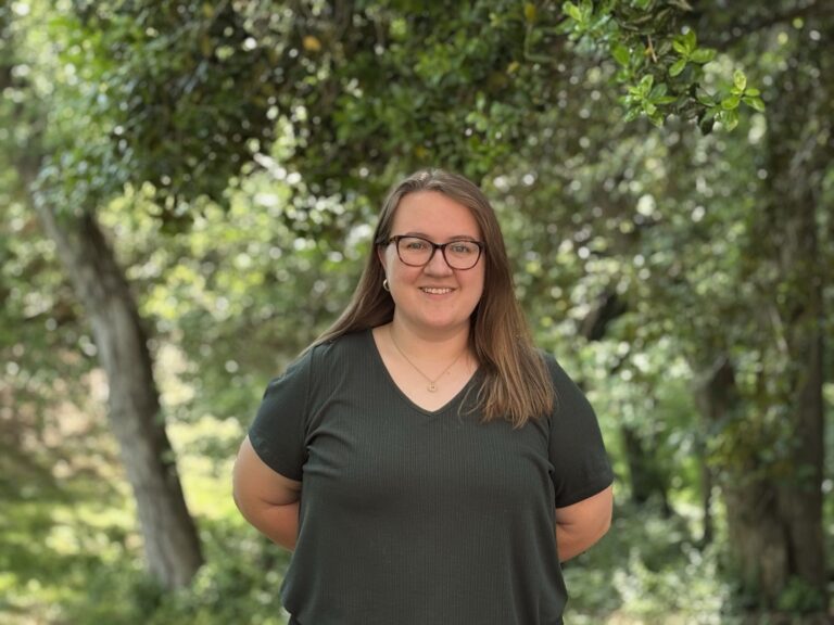 Haley Williamson in a green top smiles at the camera. Green trees are in the background.