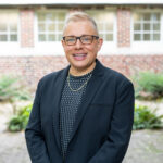 Chris Sneed stands outdoors in front of a brick building with green foliage in the background.