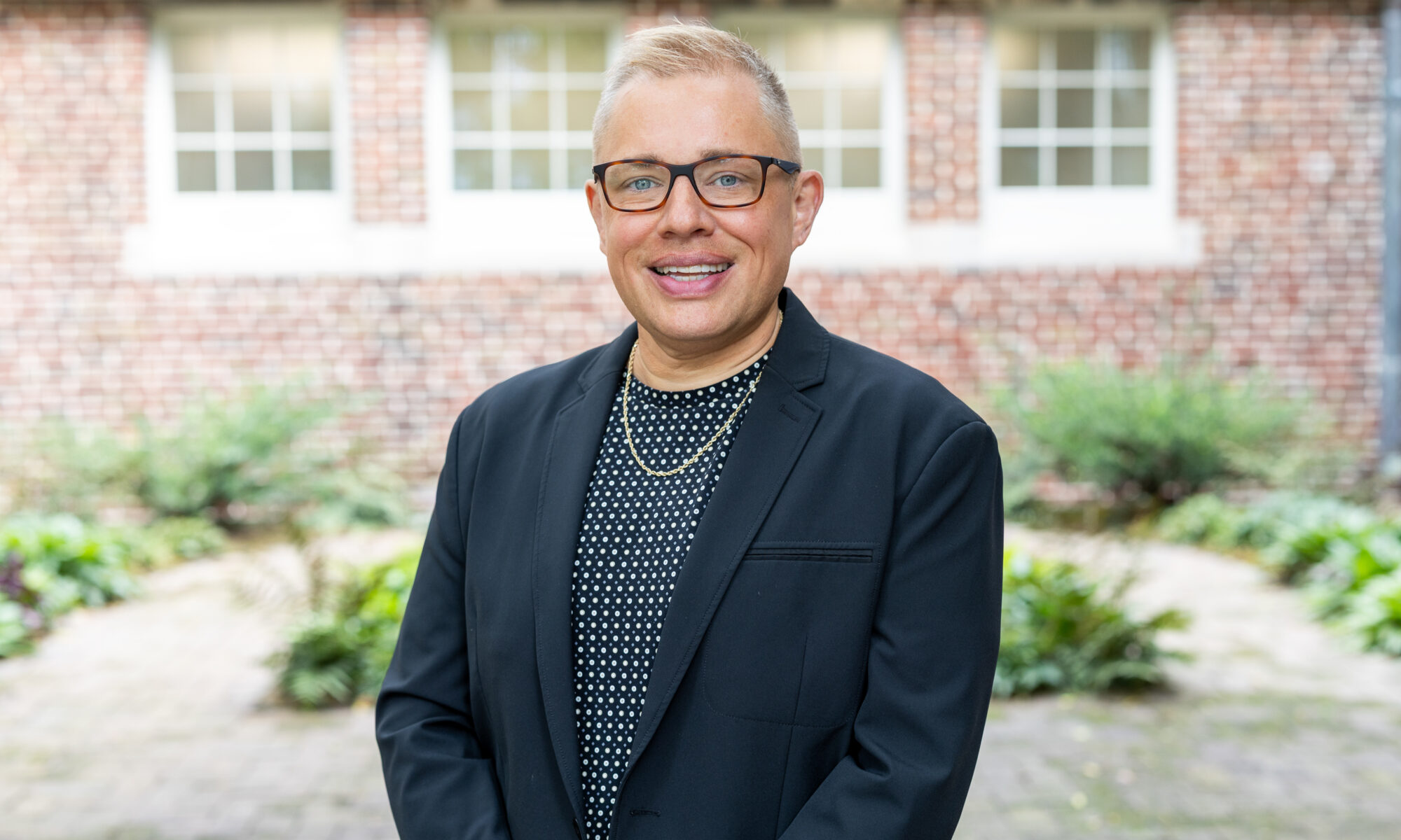 Chris Sneed stands outdoors in front of a brick building with green foliage in the background.