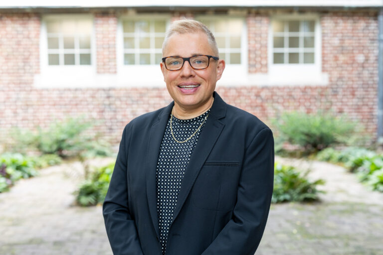 Chris Sneed stands outdoors in front of a brick building with green foliage in the background.