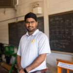Chetan Badgujar standing in a workshop-like setting. There are chalkboards in the background with writing and equipment visible in the room.