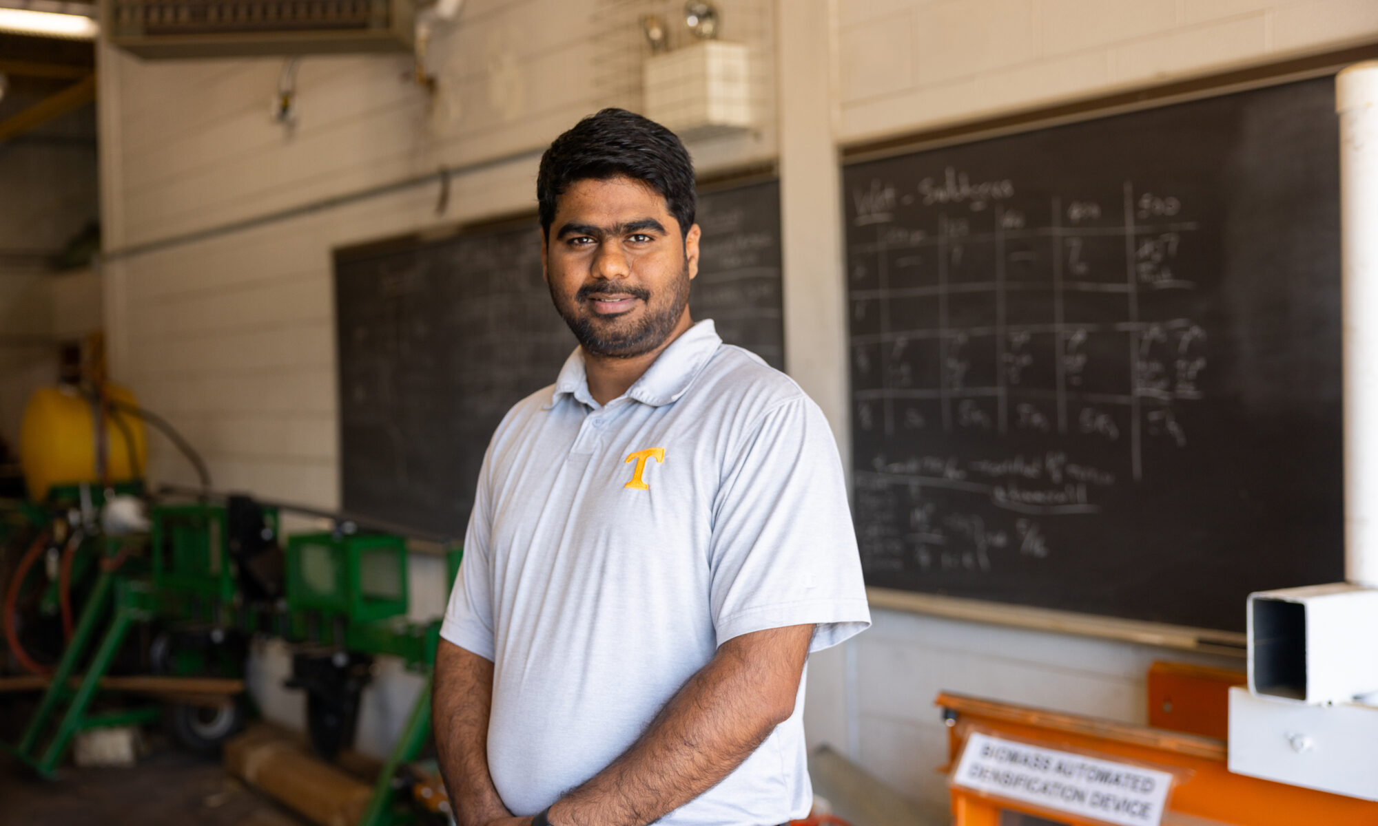 Chetan Badgujar standing in a workshop-like setting. There are chalkboards in the background with writing and equipment visible in the room.