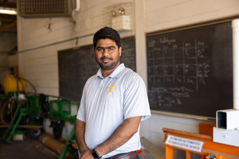 Chetan Badgujar standing in a workshop-like setting. There are chalkboards in the background with writing and equipment visible in the room.