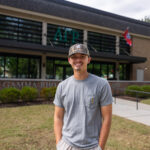 Wyatt Harvey smiling and standing in front of a building with the letters "ΑΓΡ" displayed. The building has a sign reading "Alpha Gamma Rho" and features flags above.