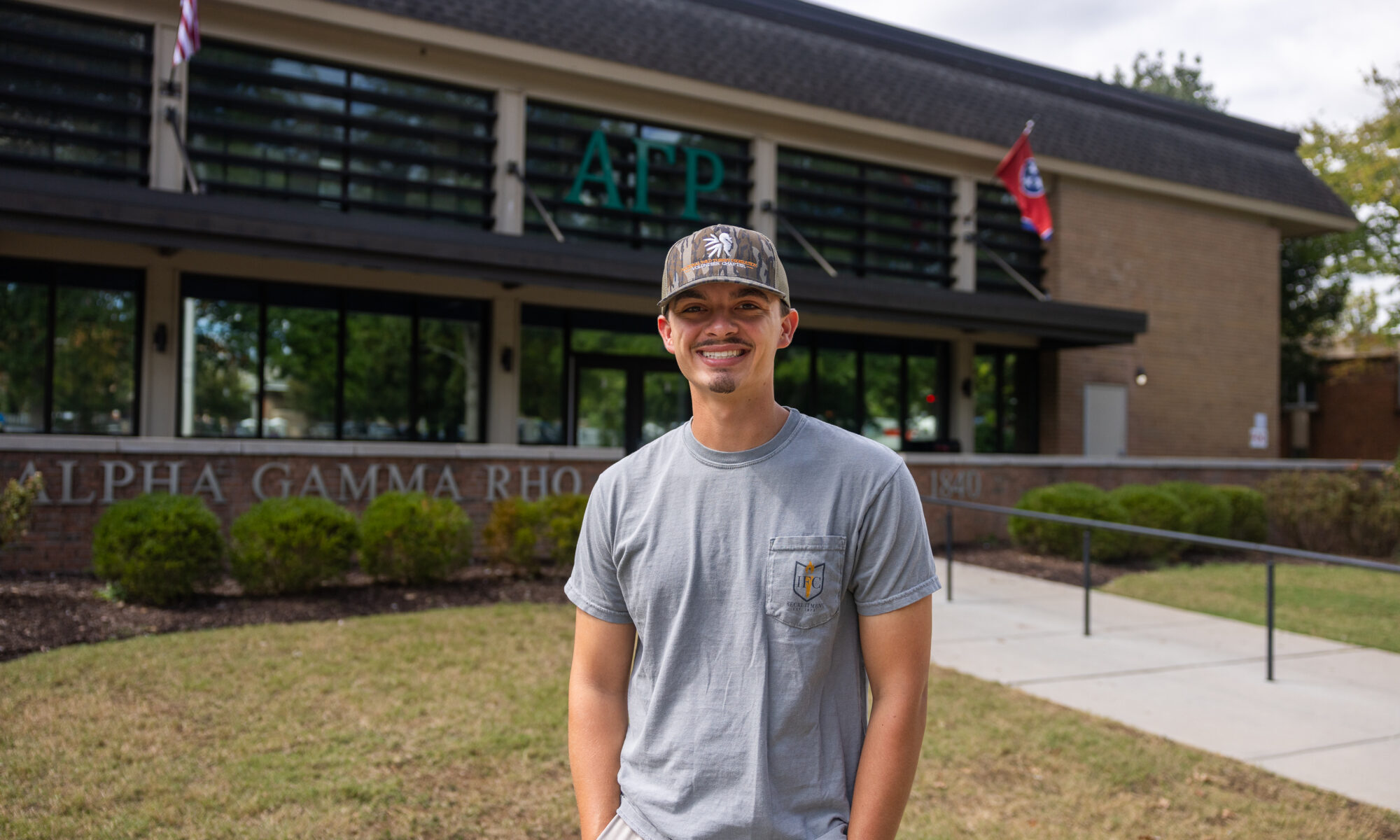Wyatt Harvey smiling and standing in front of a building with the letters "ΑΓΡ" displayed. The building has a sign reading "Alpha Gamma Rho" and features flags above.
